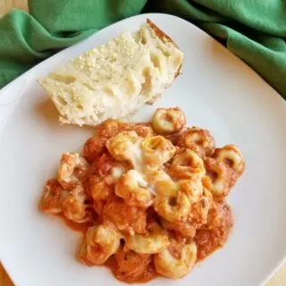 Plate of tortellini casserole with creamy tomato sauce and cheese on plate with hunk of garlic bread.