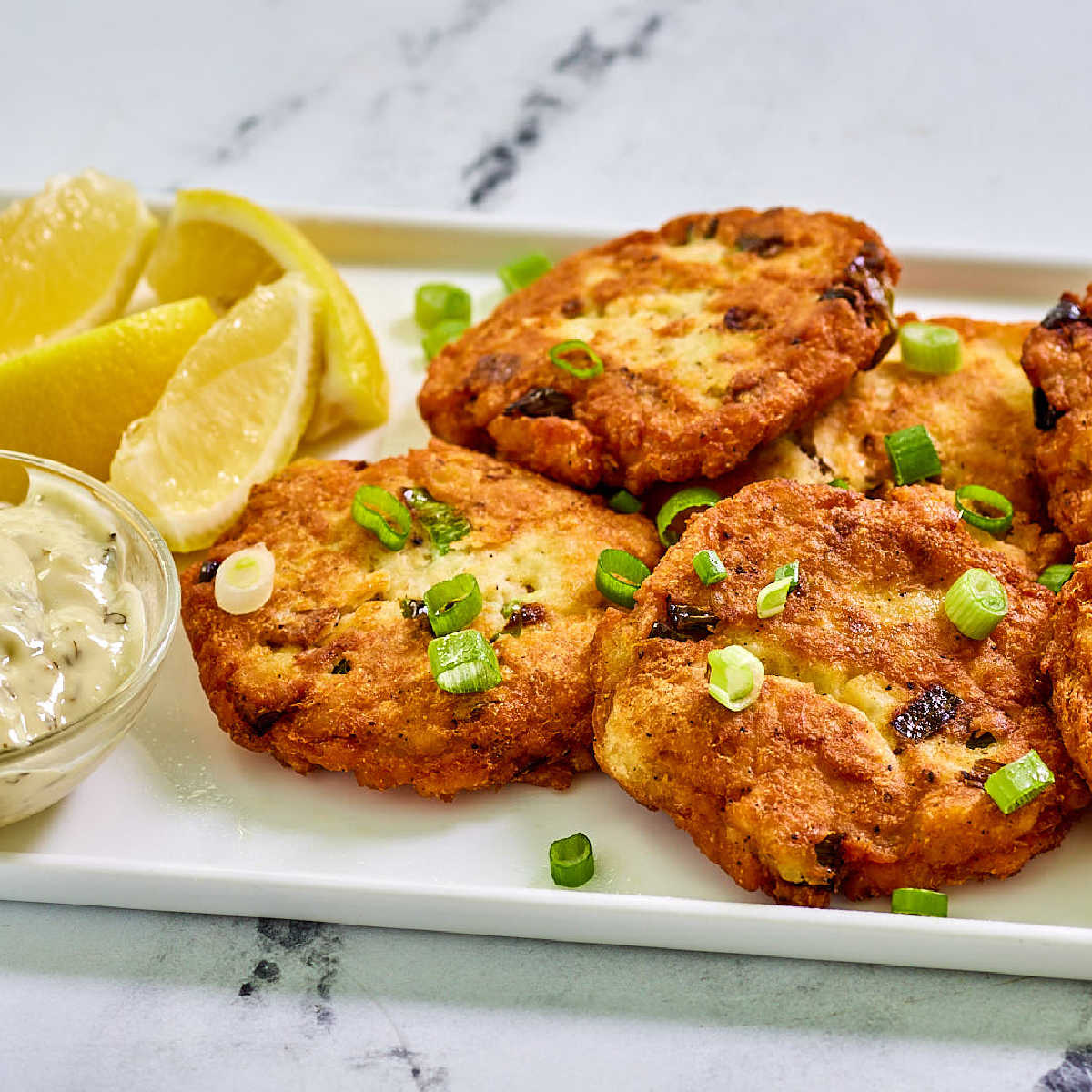 Golden salmon potato cakes topped with green onion and served on a plate with lemon wedges and tartar sauce.
