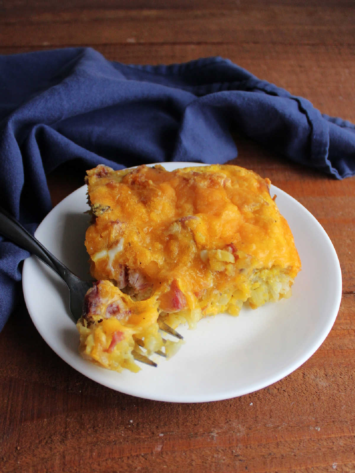 Bite of casserole on fork showing scrambled eggs, melted cheese, chunk of ham and tater tots baked inside. 