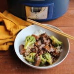 Bowl of rice topped with backstrap and broccoli with sesame seeds and chopsticks next to the crockpot it was cooked in.