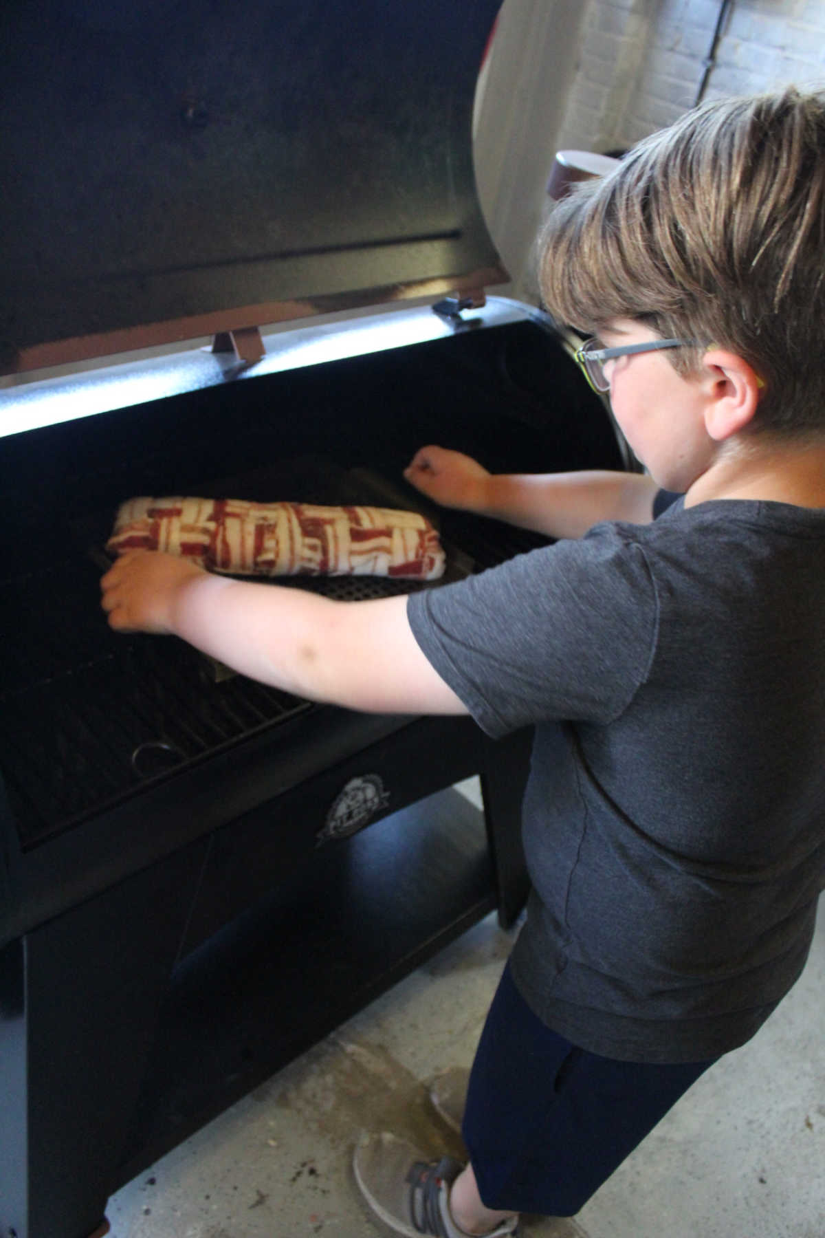 Child placing perforated grill pan with bacon explosion onto a preheated smoker grate. 