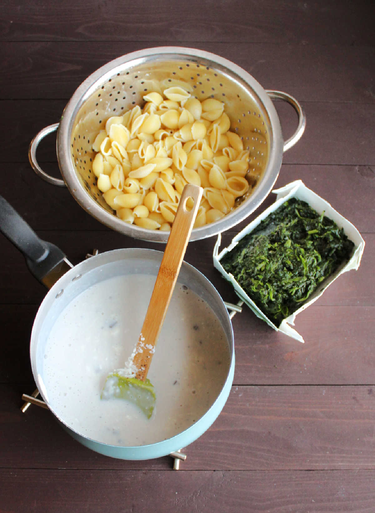 saucepan filled with white cheese sauce, colander filled with cooked shell pasta and box of defrosted chopped spinach. 
