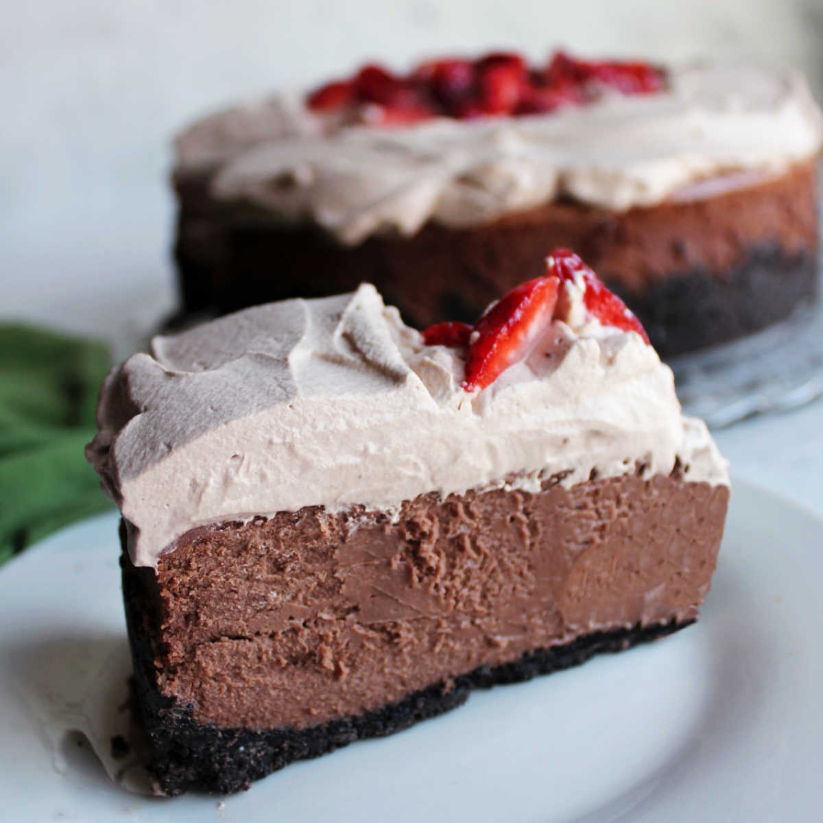 Slice of cheesecake with Oreo crust under a layer of rich chocolate cheesecake topped with light chocolate whipped cream and strawberry slices.