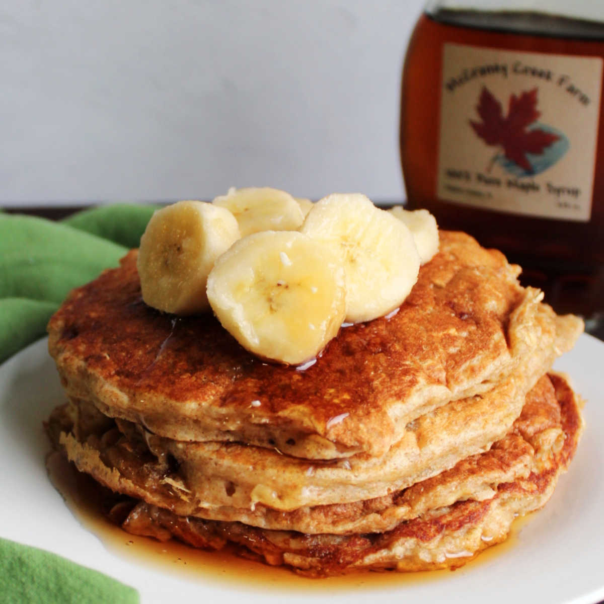Close stack of banana pancakes with oatmeal and yogurt topped with banana slices and maple syrup.