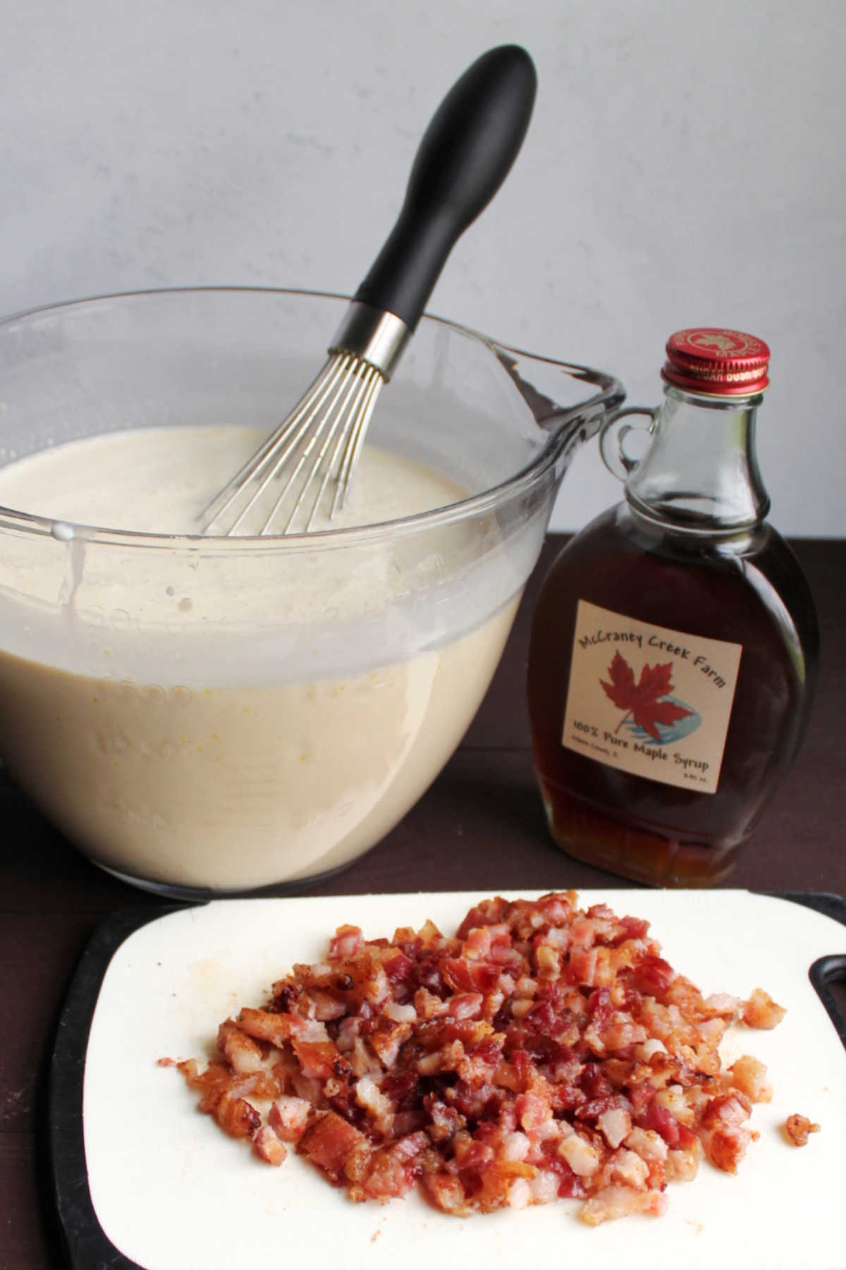 Big glass bowl of ice cream base next to a pile of chopped bacon and bottle of maple syrup.