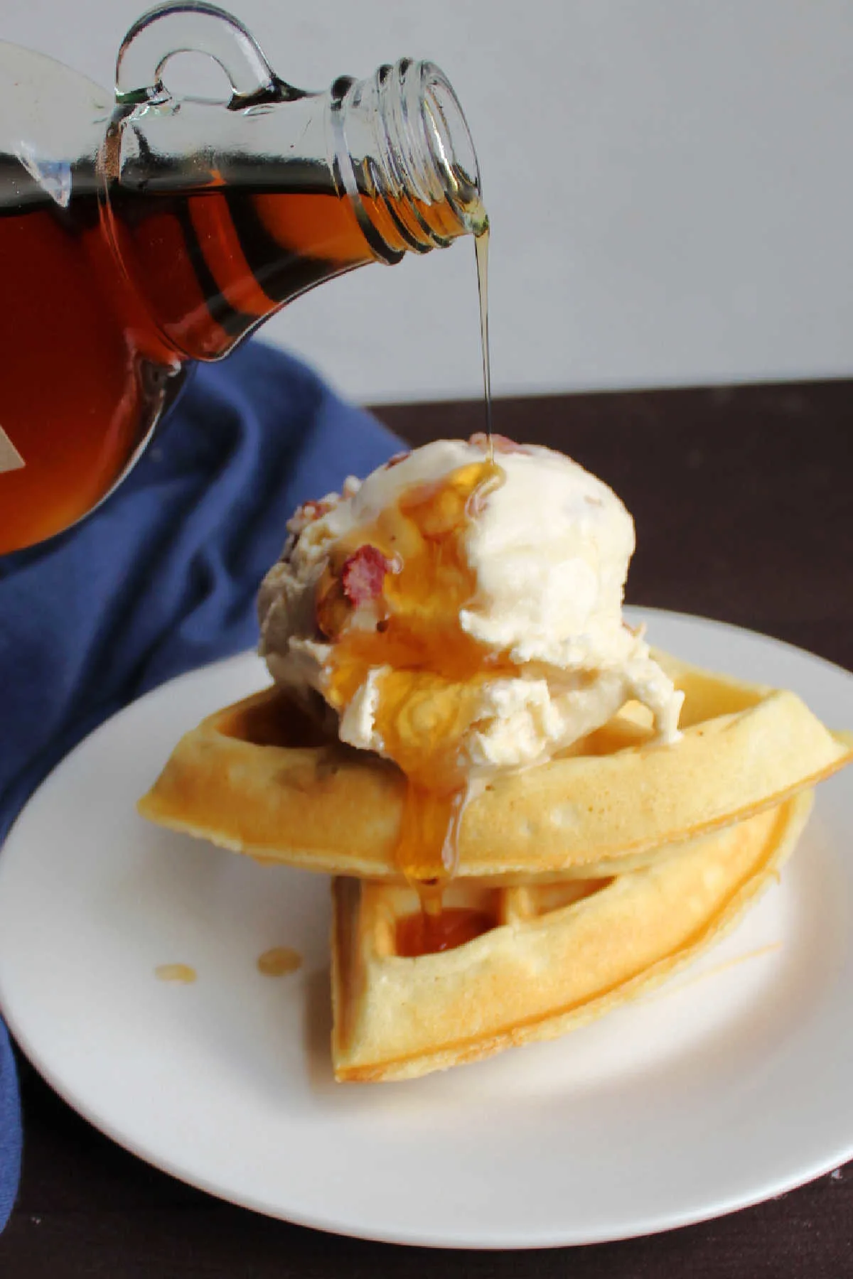 Pouring maple syrup over scoop of maple bacon ice cream perched on top of a couple pieces of Belgian waffle.
