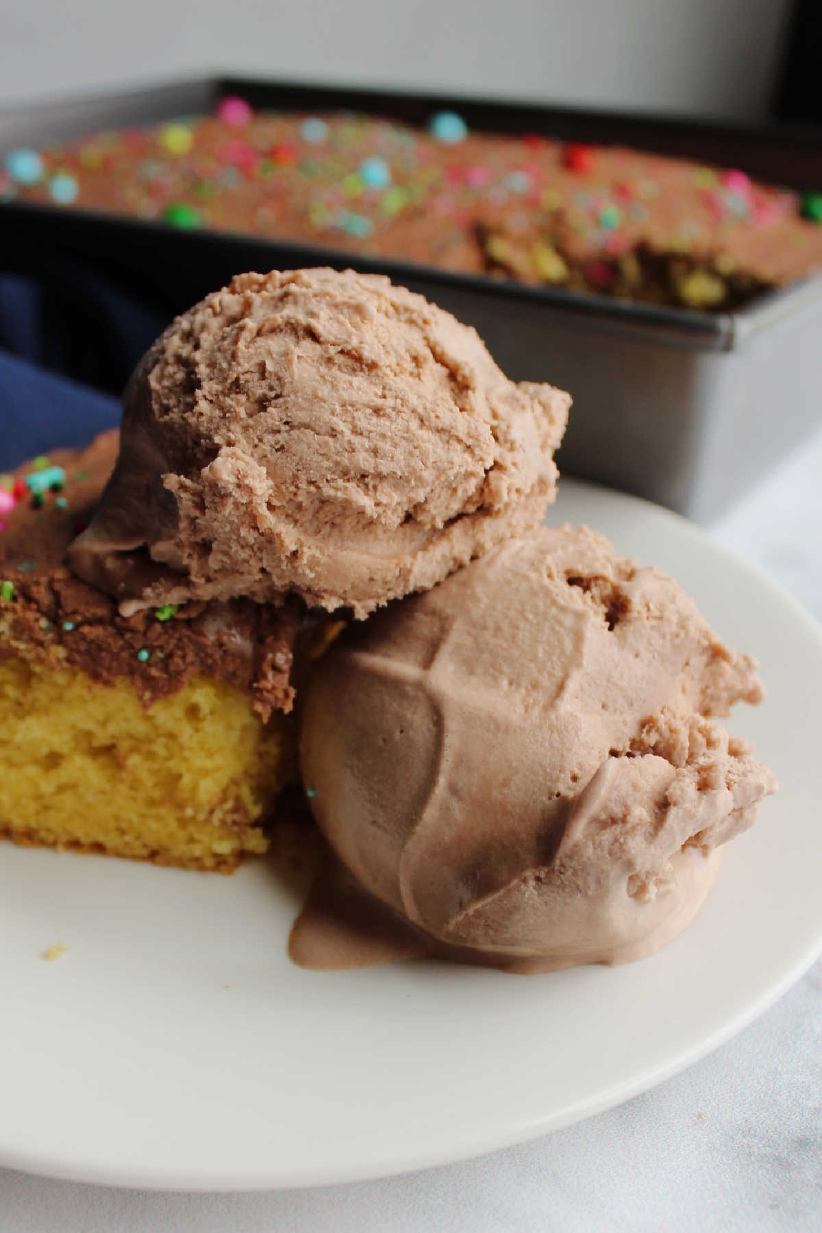 Two scoops of brownie batter ice cream on plate with cake with more cake in background.