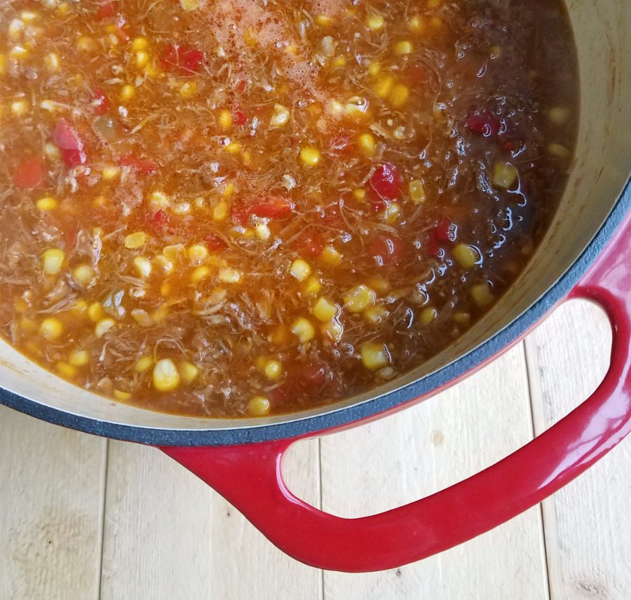 Enameled cast iron dutch oven filled with tomato based brunswick stew with pulled pork, chicken, brisket, and corn in it.