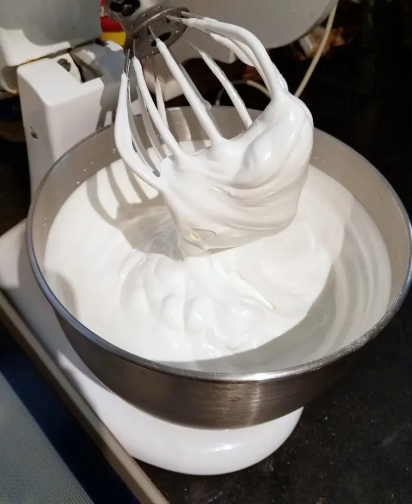 Stand mixer filled with glossy meringue with bowlful of meringue underneath it. 