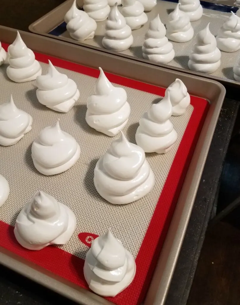 Piped swirls of glossy meringue on baking sheets ready to go in the oven.