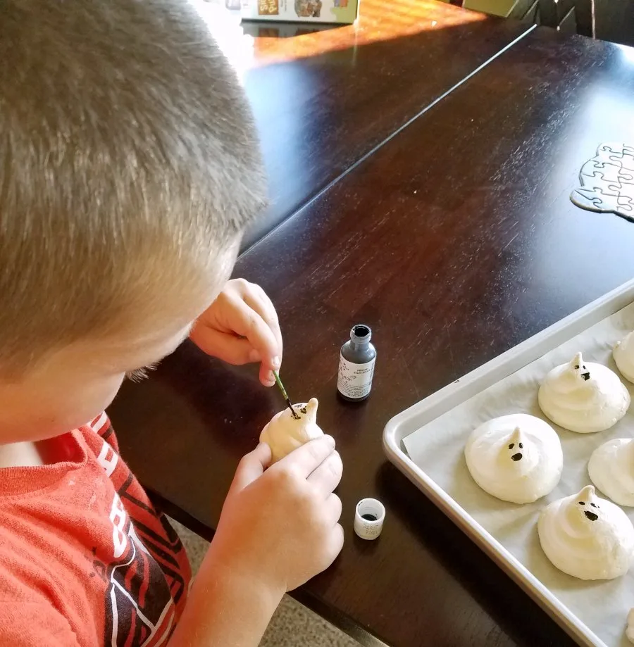 Little Dude using a toothpick to paint black food coloring gel faces on crisp meringue ghost cookies.