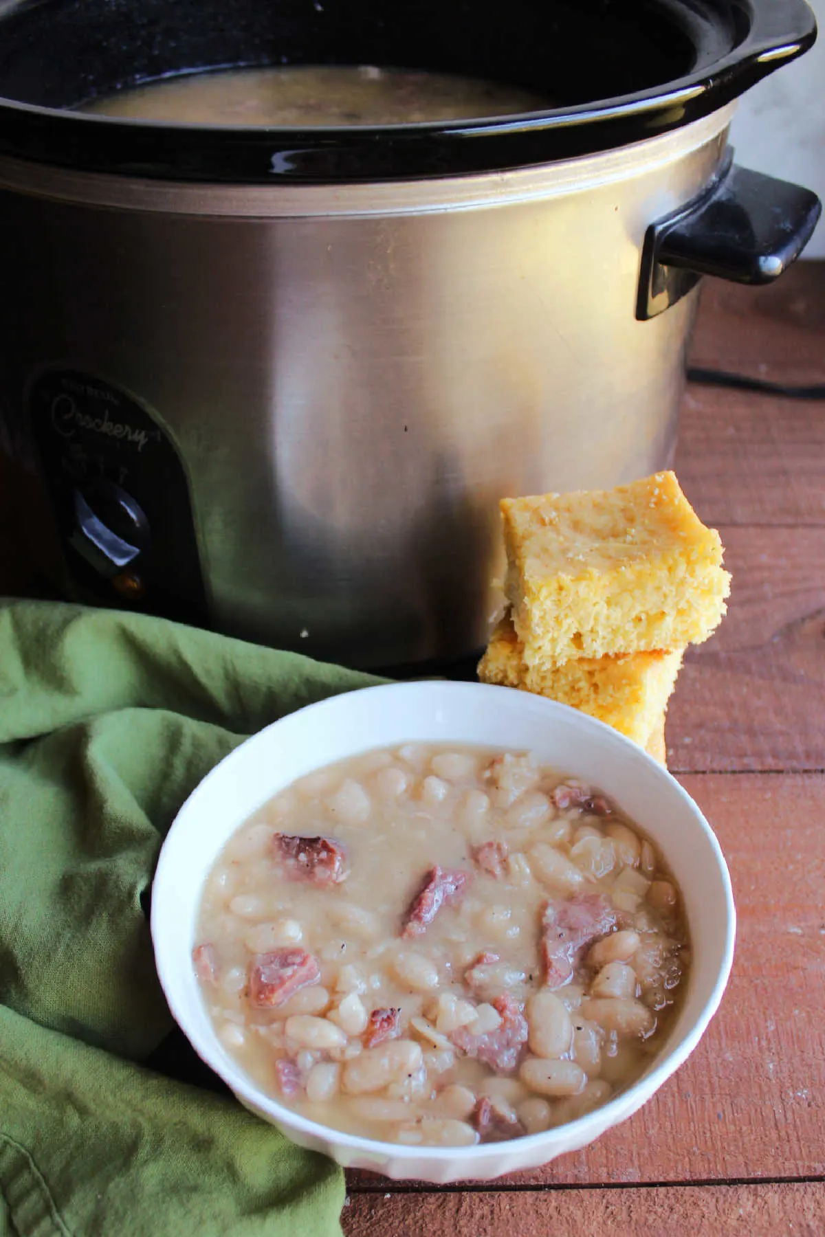 Bowl of ham and beans in front of slow cooker and a stack of cornbread.