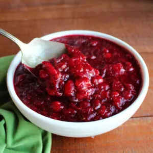 Antique berry spoon scooping out freshly made orange cranberry sauce with chunks of cranberries and a deep red color from a white serving bowl.