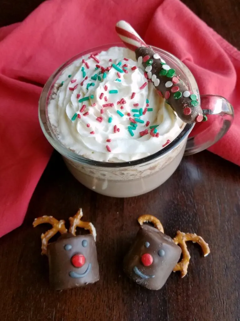 Glass mug filled with creamy slow cooker hot cocoa topped with whipped cream and sprinkles garnished with a chocolate dipped peppermint stick with two rudolph marshmallows nearby.