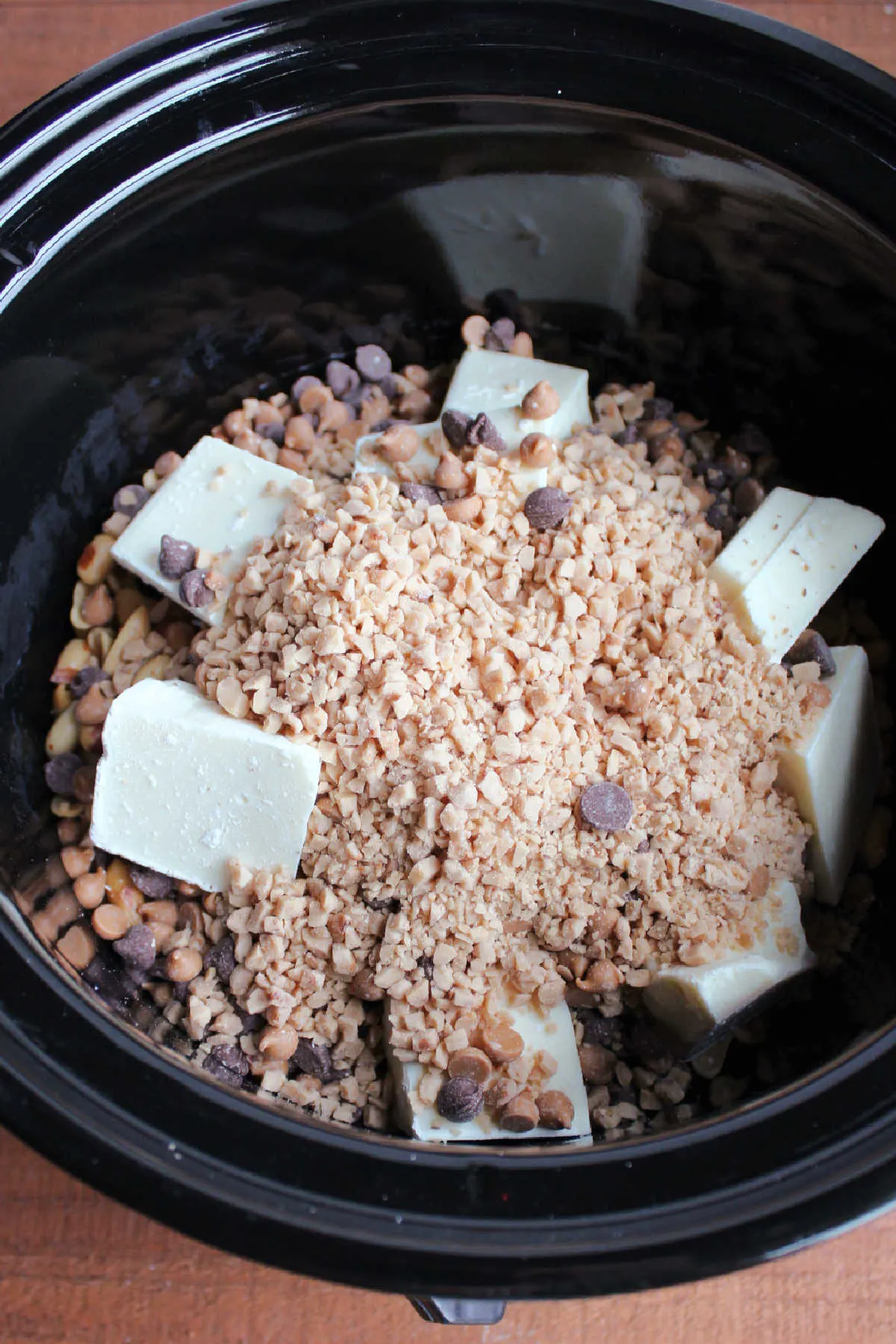 Slow cooker filled with peanuts, almond bark, chocolate chips and toffee bits ready to cook.