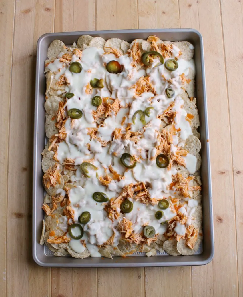 Sheet pan filled with buffalo chicken nachos topped with white cheese sauce and jalapenos.
