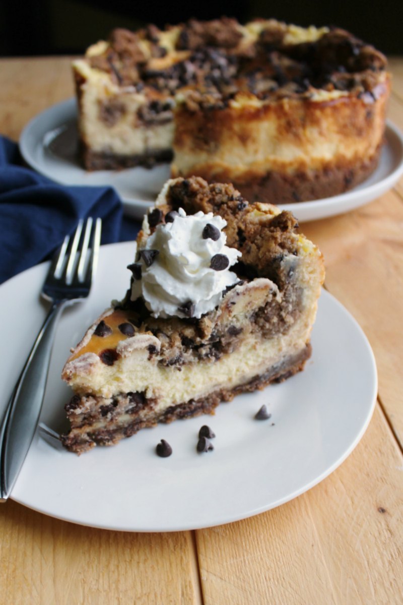 slice of chocolate chip cookie dough cheesecake with chocolate chip cookie crust topped with whipped cream and chocolate chips in front of remaining cheesecake. 