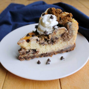 Slice of cookie dough cheesecake with chocolate chip cookie crust, creamy cheesecake center, and bits of chocolate chip cookie dough baked on top finished with some whipped cream and extra chocolate chips.
