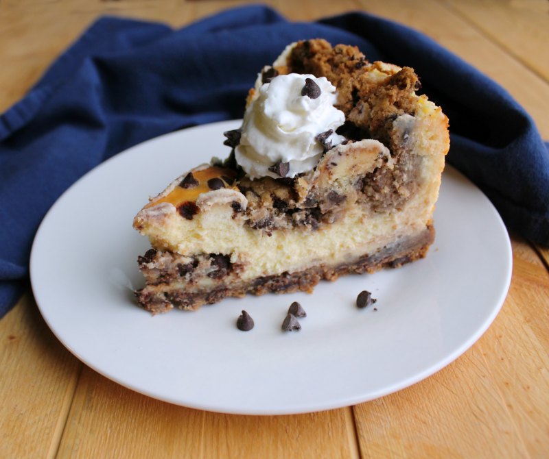 Slice of chocolate chip cookie dough cheesecake on dessert plate with navy napkin in the background.