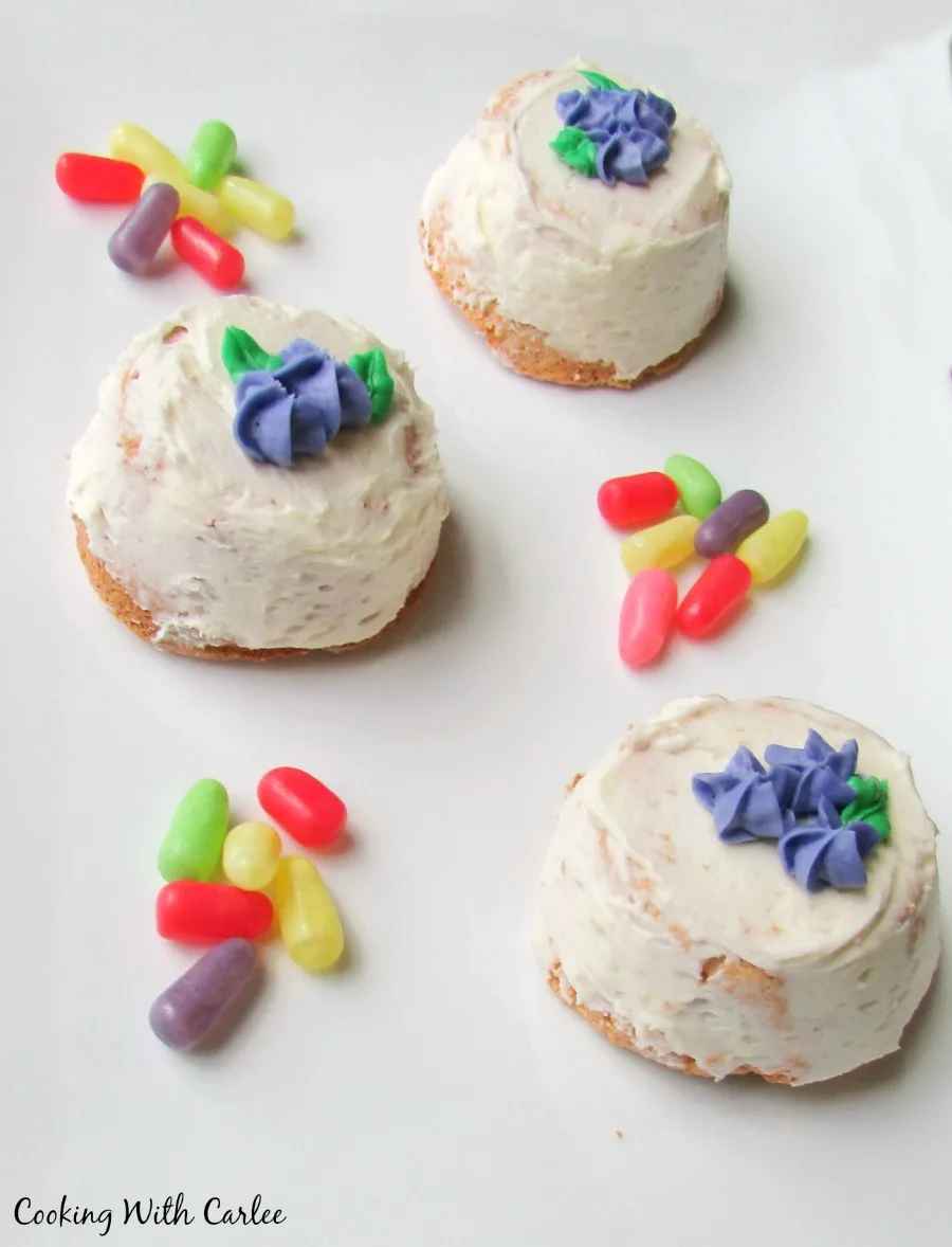 mini angel food cakes with white frosting and sides and purple frosting flowers on top, surrounded with bright colored candy.