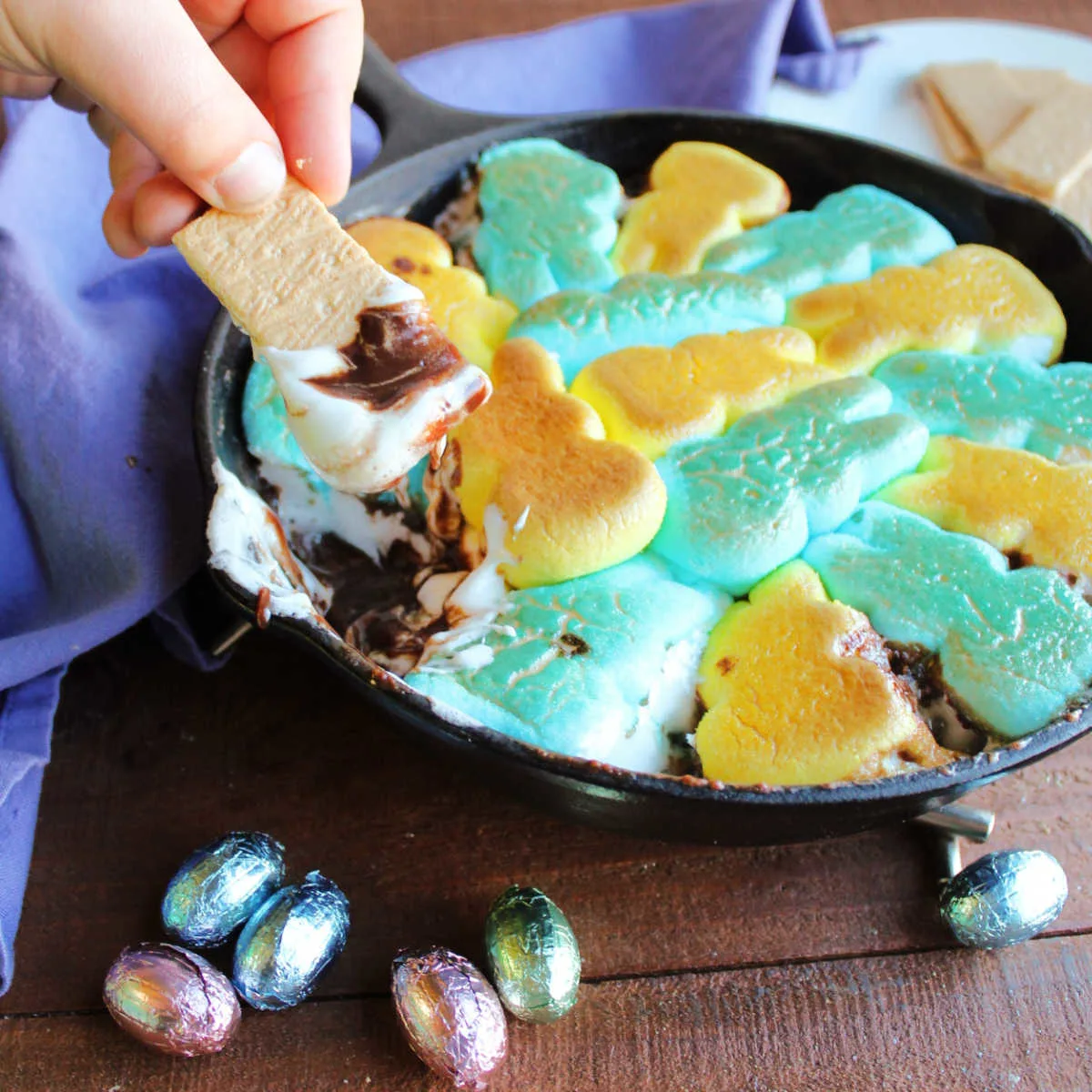 Child's hand holding piece of graham cracker dipped into smore dip with gooey melted Peeps on top of melted chocolate mixture.