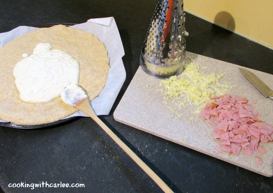 pizza crust with bechamel on it next to cutting board with ham and cheese.
