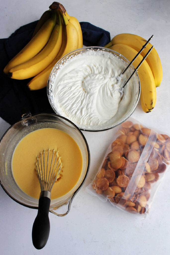 Bowl of pudding mixture, bowl of fresh whipped cream, bunches of bananas and vanilla wafers ready to be made into banana pudding.