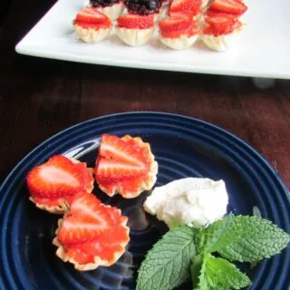 plate of mini strawberry tarts in front of platter with strawberry and blueberry tarts arranged like an American flag.
