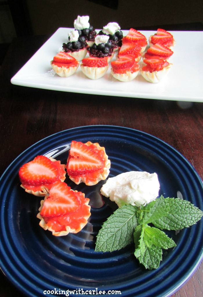 plate of mini strawberry tarts in front of platter with strawberry and blueberry tarts arranged like an American flag.