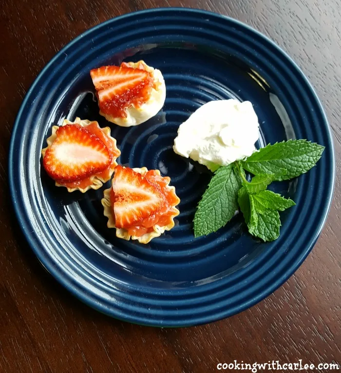 Plate with 3 strawberry tartlets, a dollop of whipped cream and a sprig of mint.
