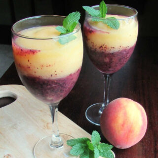 2 wine glasses with layers of peach and blueberry wine slushy next to peaches, blueberries and fresh mint leaves