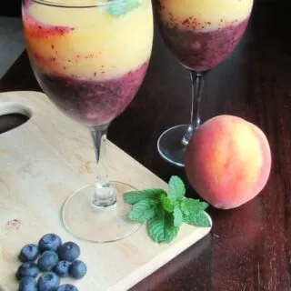 Two wine glasses with layer of blueberry wine slush in the bottom and peach wine slush on the top garnished with mint leaves.