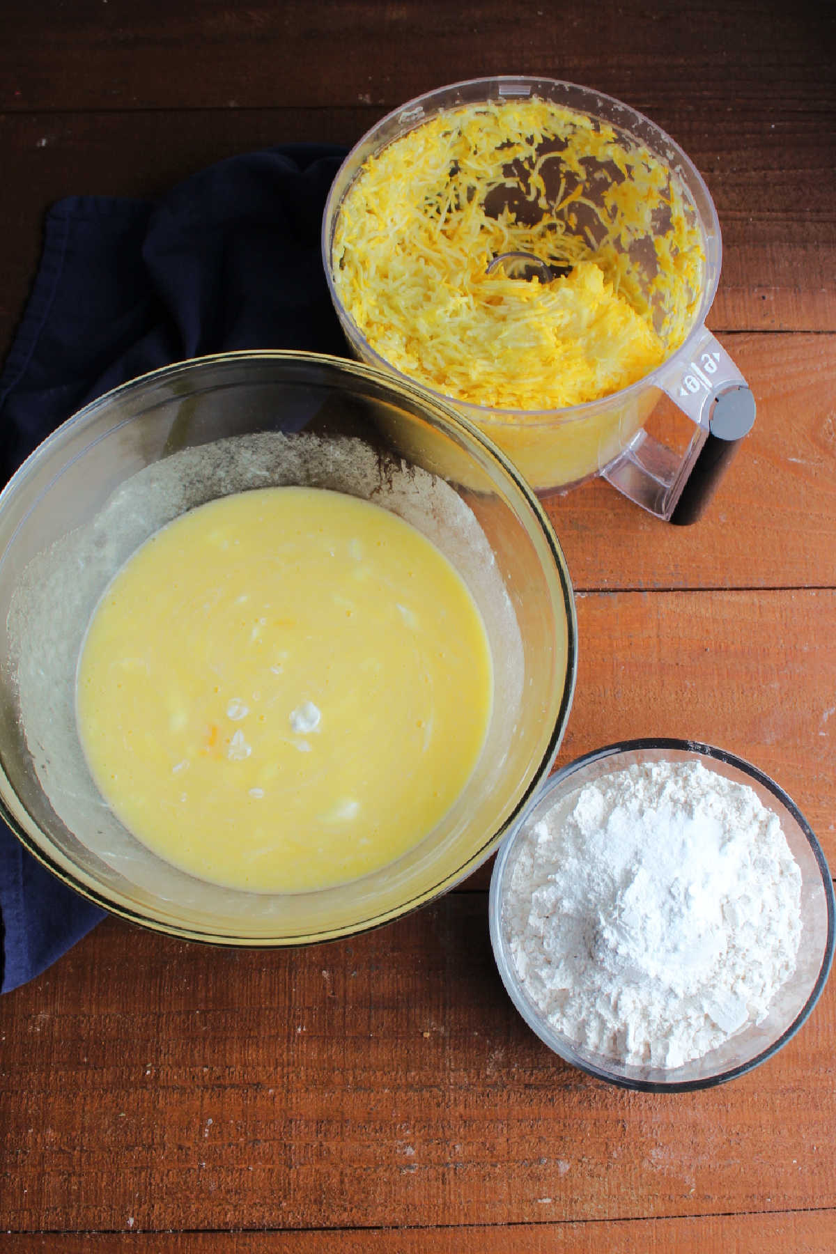 Bowl of dry ingredients, next to mixing bowl of wet ingredients, with a food processor filled with shredded yellow squash nearby.