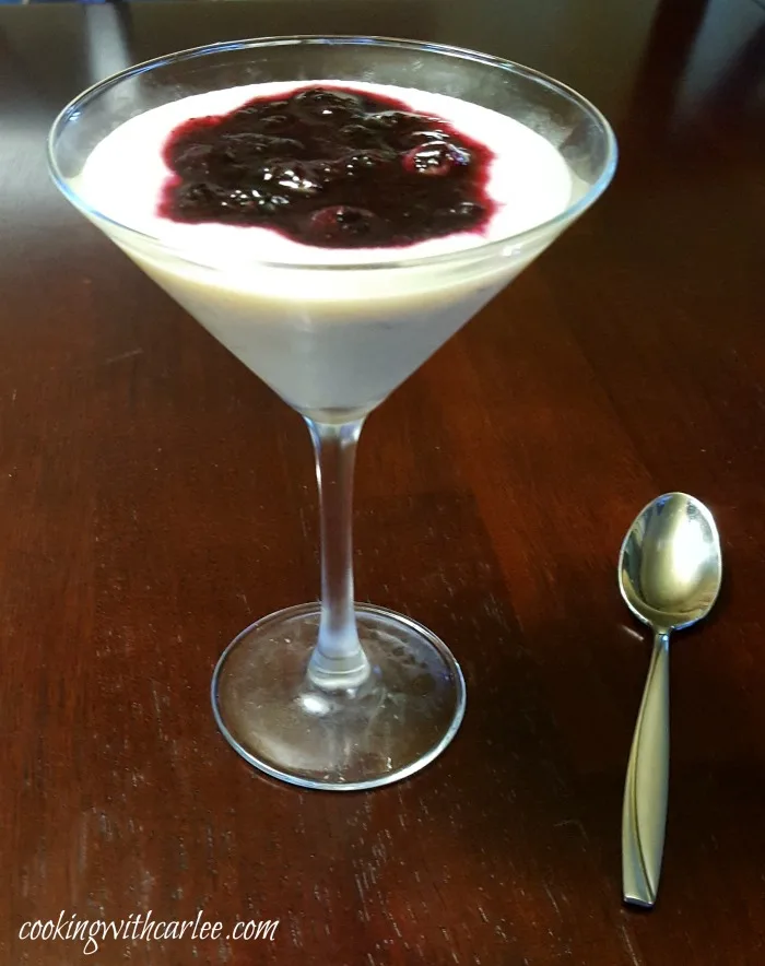 martini glass filled with creamy cornmeal budino and blueberry sauce with a spoon nearby ready to eat.