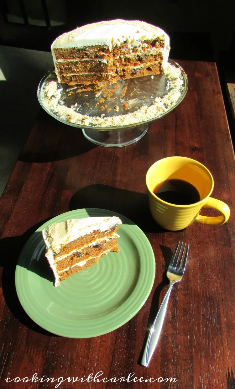 three layer carrot cake with raisins and cream cheese frosting on cake plate with slice on plate next to cup of coffee.