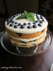 3 layer blueberry cake topped with lemon cream cheese frosting, fresh berries and a sprig of fresh mint on top.