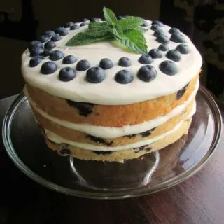 3 layer blueberry cake topped with lemon cream cheese frosting, fresh berries and a sprig of fresh mint on top.