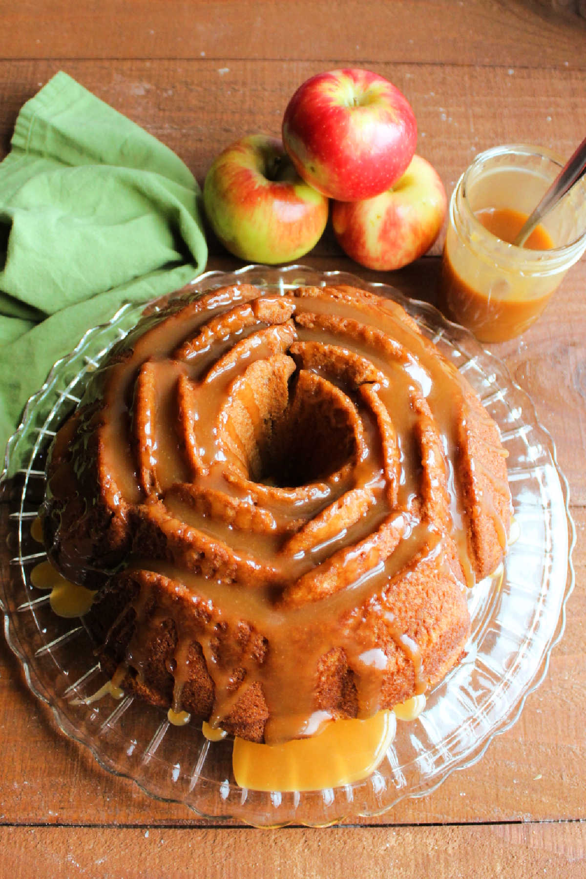 Whole flower shaped apple cider bundt cake with apples and a jar of caramel sauce in the background.