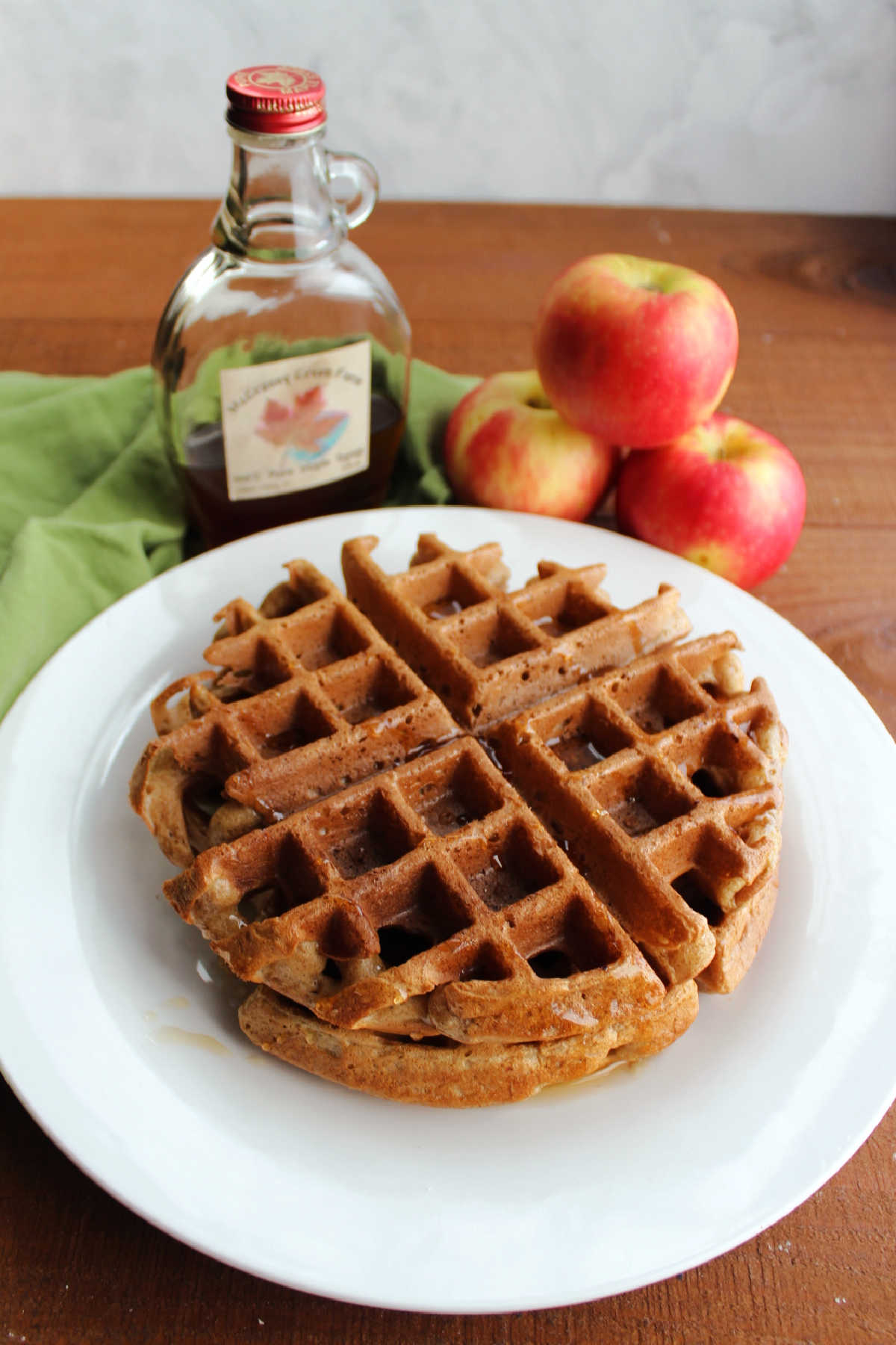 Stack of apple cider waffles with bottle of maple syrup and apples.