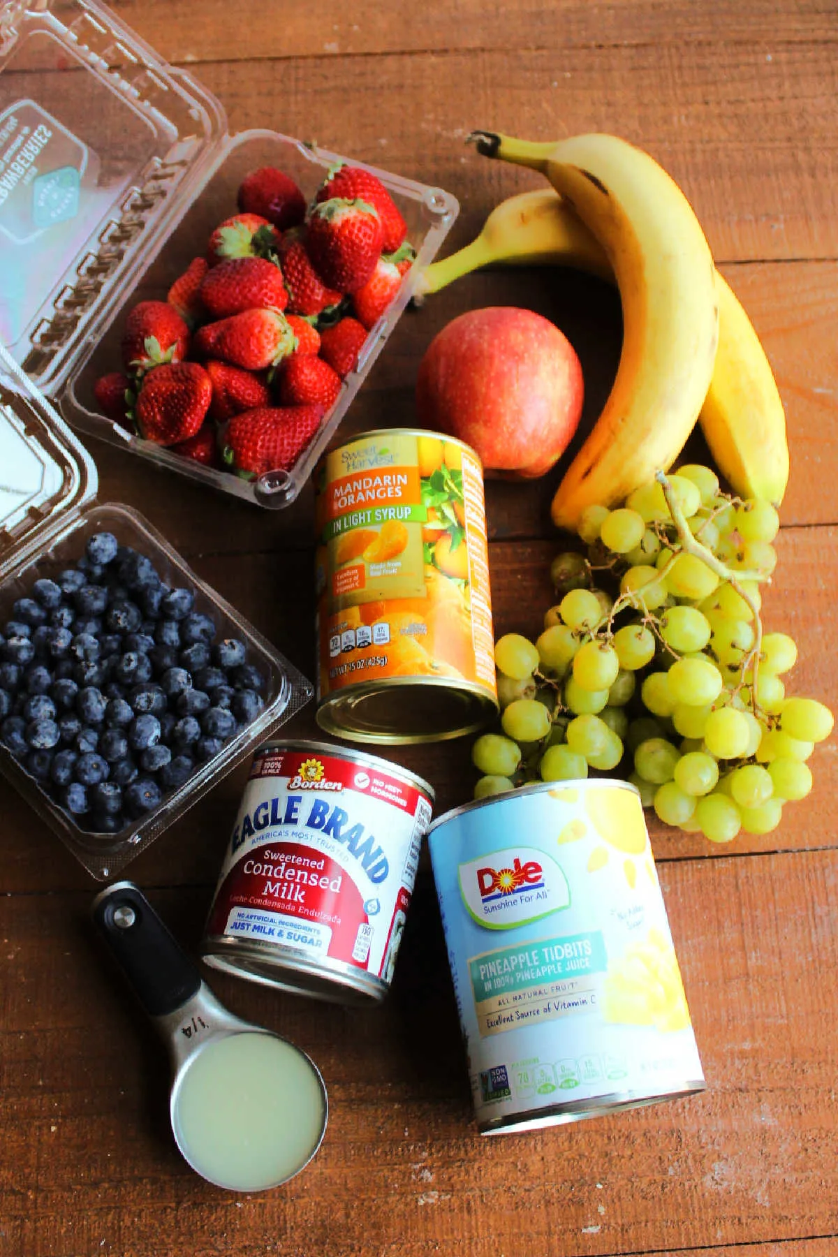 Ingredients including fresh bananas, strawberries, blueberries, grapes, apple and canned oranges, pineapple, condensed milk and lemon juice ready to be made into condensed milk fruit salad.