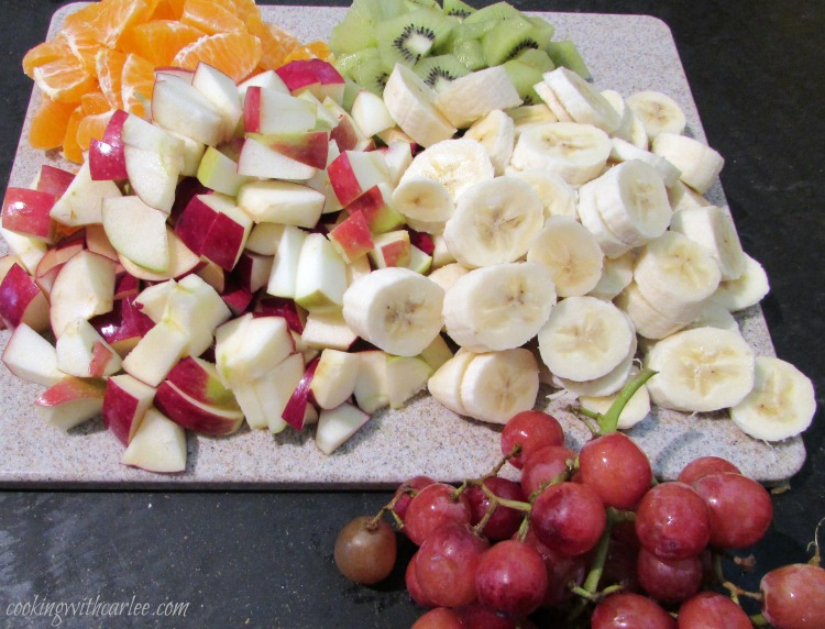Cutting board filled with sliced bananas, kiwi, chopped apples and oranges.