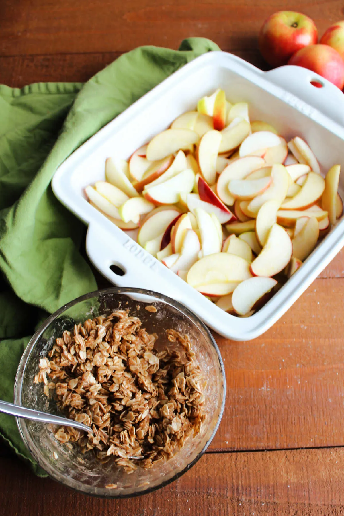 Sliced apples in square baking dish with small bowl of oat topping nearby.