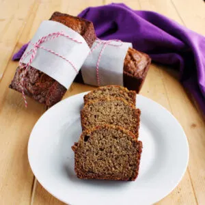 Slices of mini whole wheat banana bread loaf on plate with mini loaves wrapped in parchment paper and bakers twine in the background.