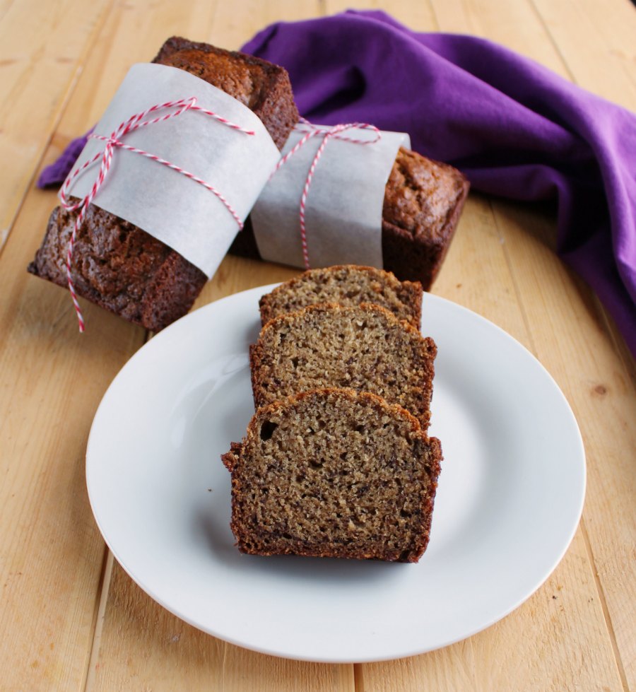 Slices of mini whole wheat banana bread loaf on plate with mini loaves wrapped in parchment paper and bakers twine in the background. 