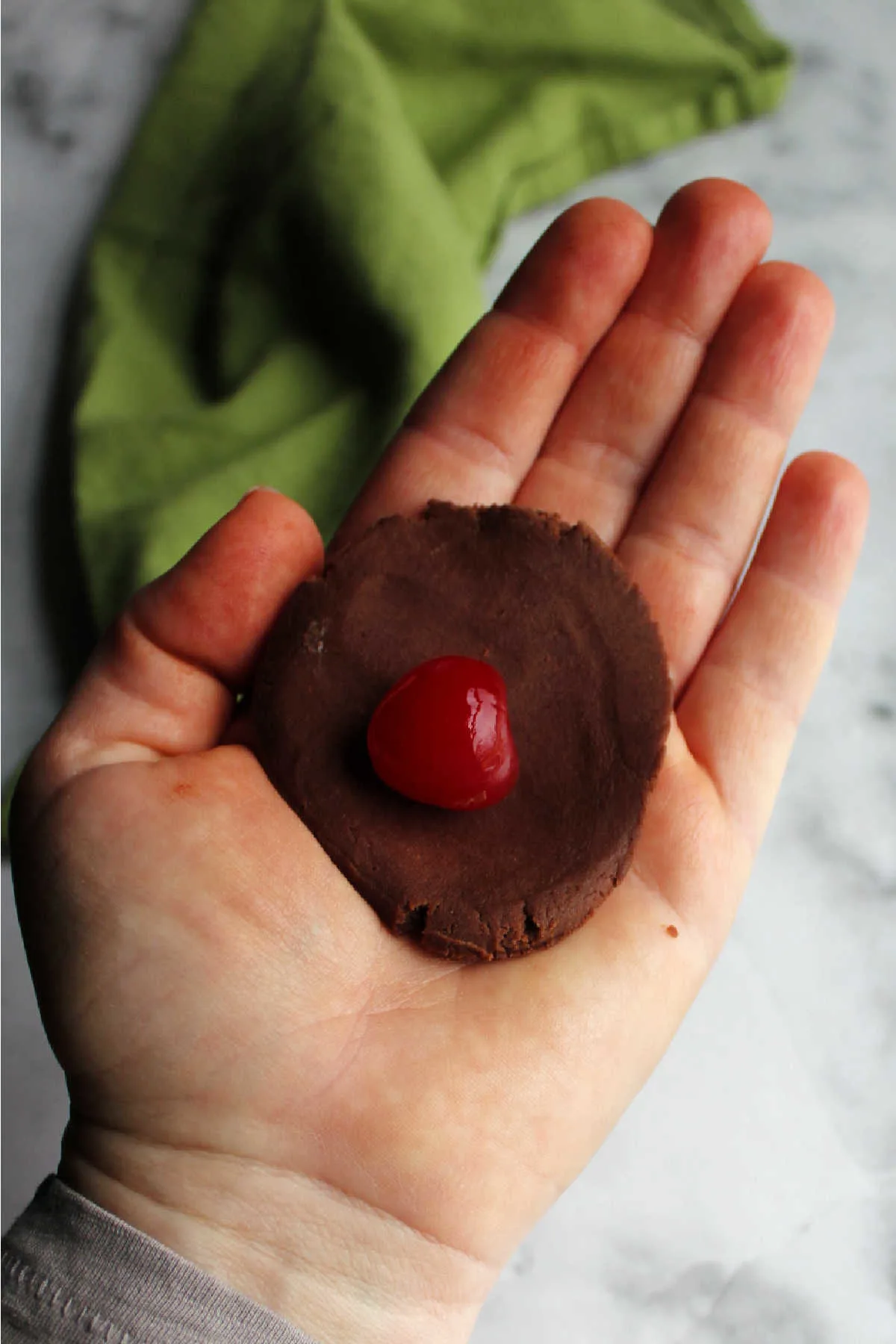 Hand with a circle of chocolate cookie dough and a cherry in the center ready to wrap the dough around the cherry.