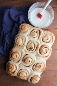 Freshly baked potato cinnamon rolls with golden brown tops next to a mixing bowl with vanilla icing inside.