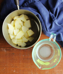 Saucepan with cooked chunks of potatoes in it next to a measuring cup with water from the cooked potatoes and sugar inside.