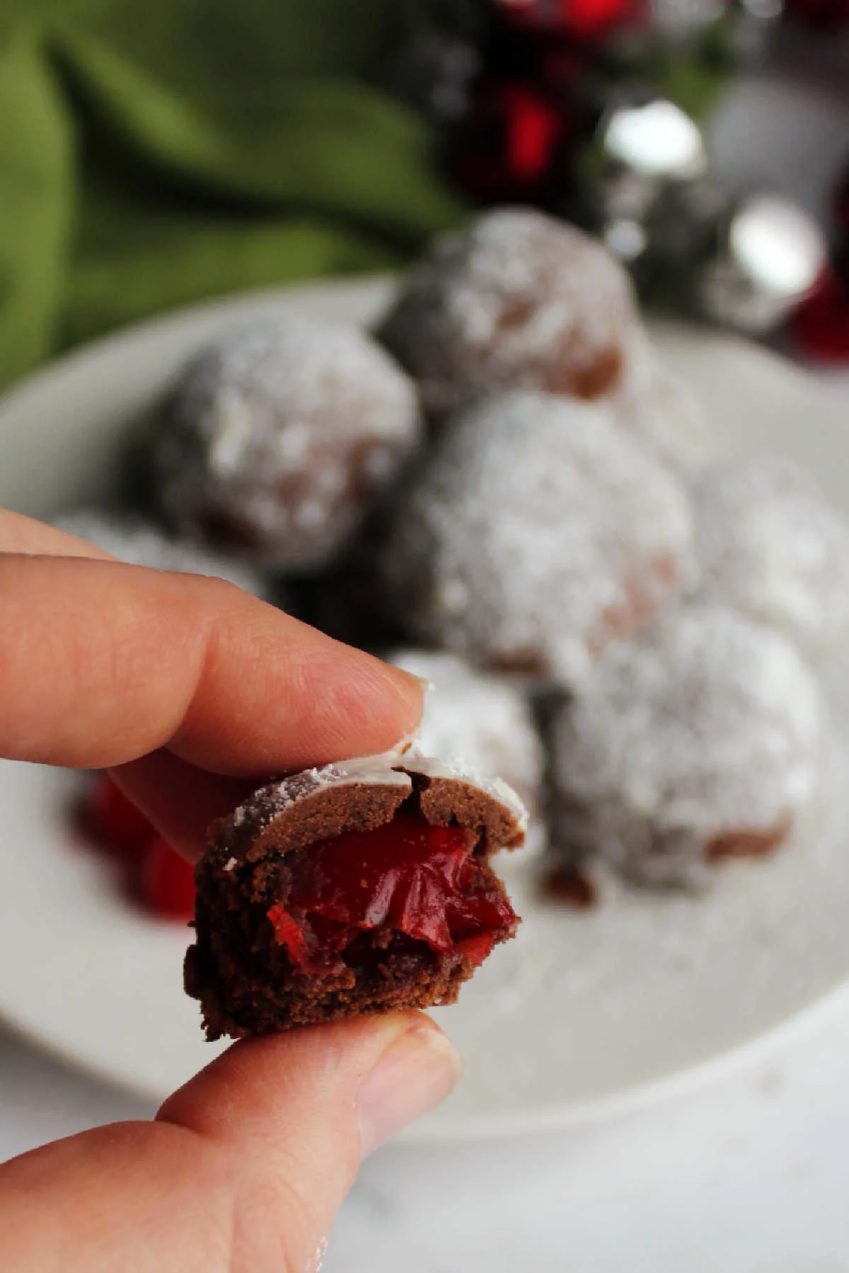 Hand holding half of a chocolate cookie with powdered sugar on the outside and a cherry on the inside. 