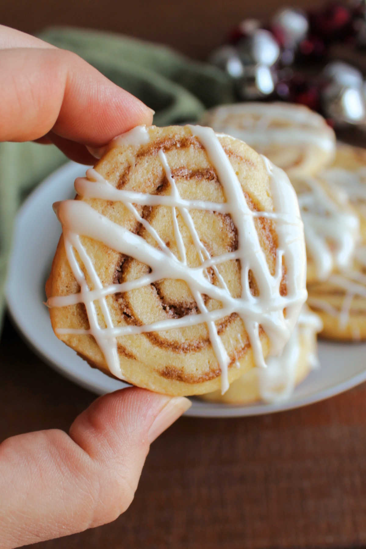 Hand holding a cinnamon roll cookie with swirls of cinnamon filling baked inside and cream cheese icing drizzled over the top.