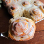 Spatula lifting large golden brown potato cinnamon roll topped with vanilla icing out of pan of freshly baked cinnamon rolls.